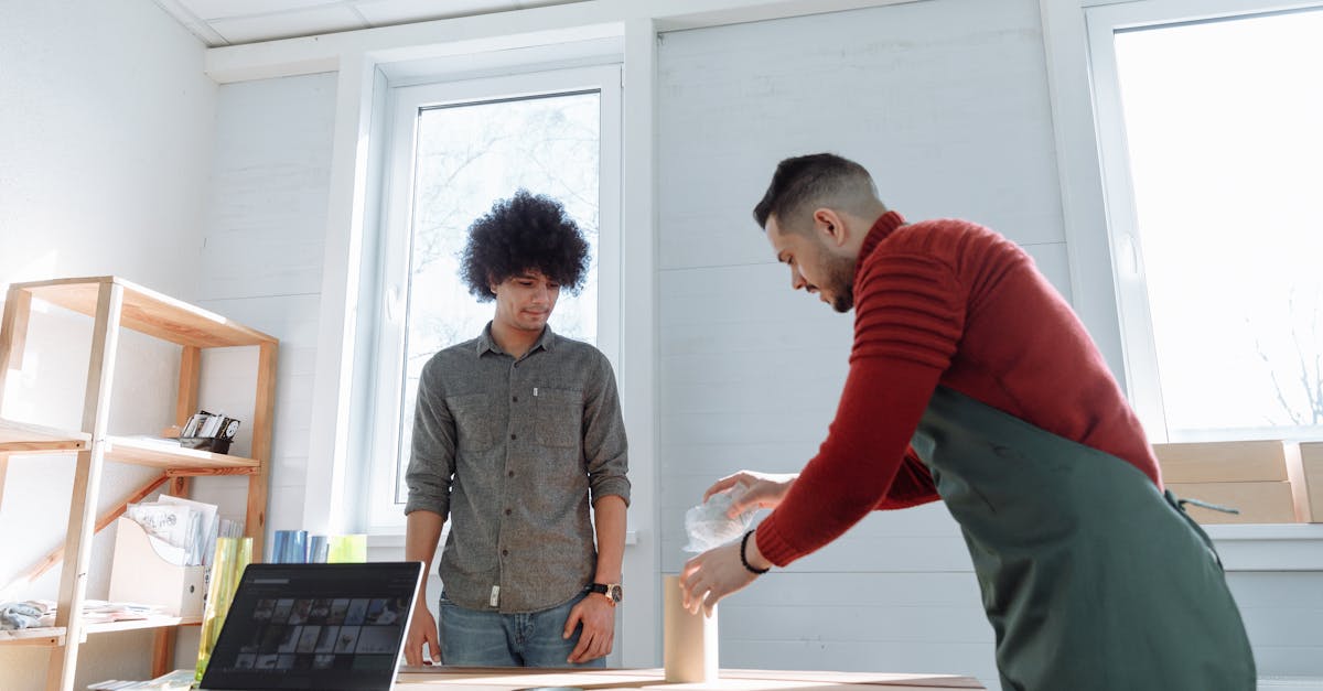 Two adults collaborating at a standing desk in a bright office with a laptop and packaging materials.