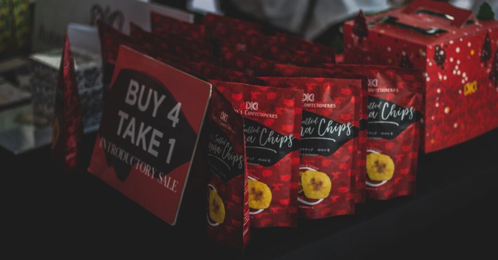 Packaged banana chips displayed with a promotional 'Buy 4 Take 1' sign at an indoor sale event.