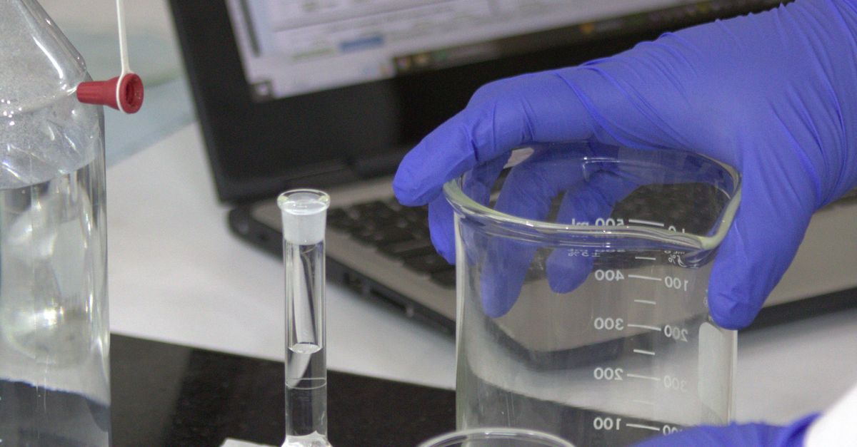 Scientist in gloves with beaker and laptop in a lab, conducting experiments.