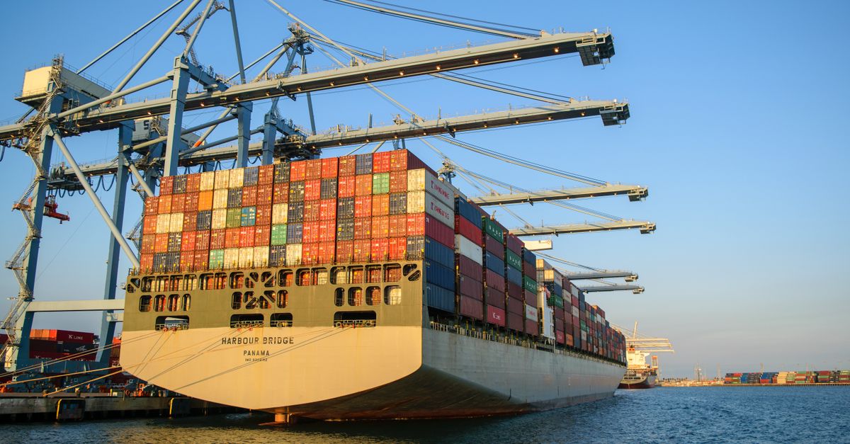 A large container ship at an industrial harbor with cranes under a clear blue sky.