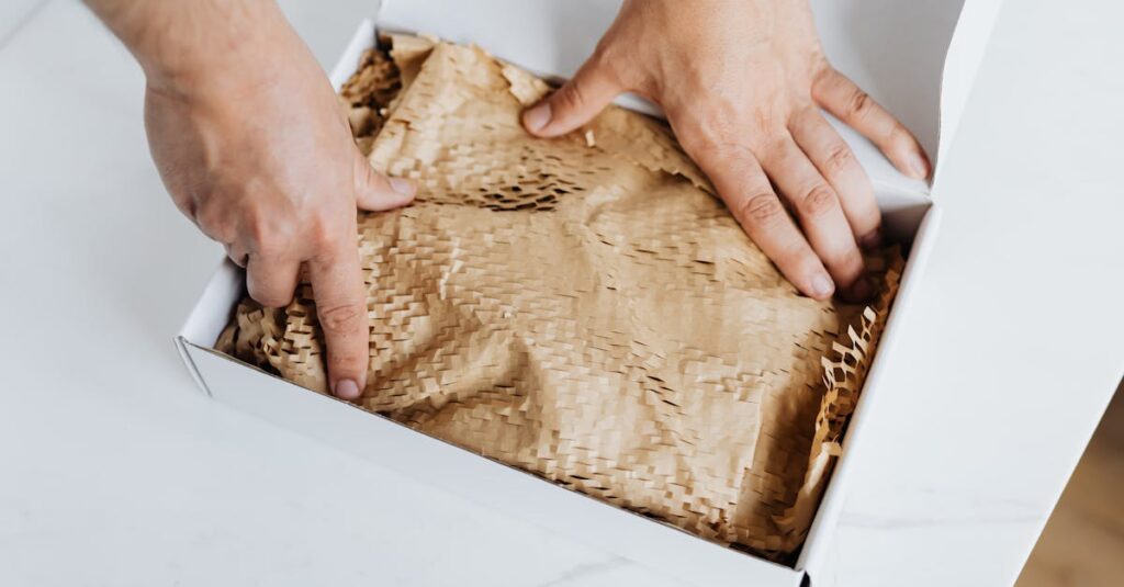 From above crop adult male packing ordered product or gift in white cardboard box with craft paper for safety postal delivery at white marble table
