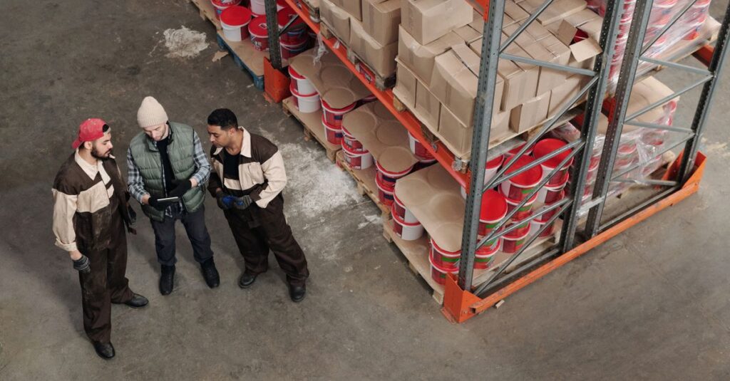 Three workers discussing logistics in a warehouse with shelves and packages.
