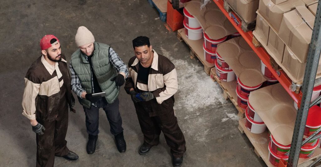 Three workers in a warehouse discussing inventory management near shelves with boxes and paint cans.