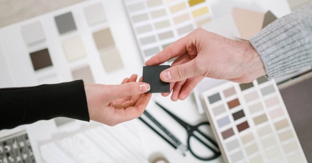 Two hands exchange a color sample during a collaborative design meeting with various palettes.