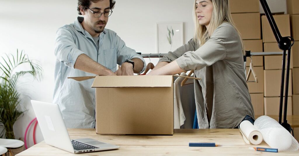 Man and woman packing e-commerce orders at startup business with laptop and boxes indoors.