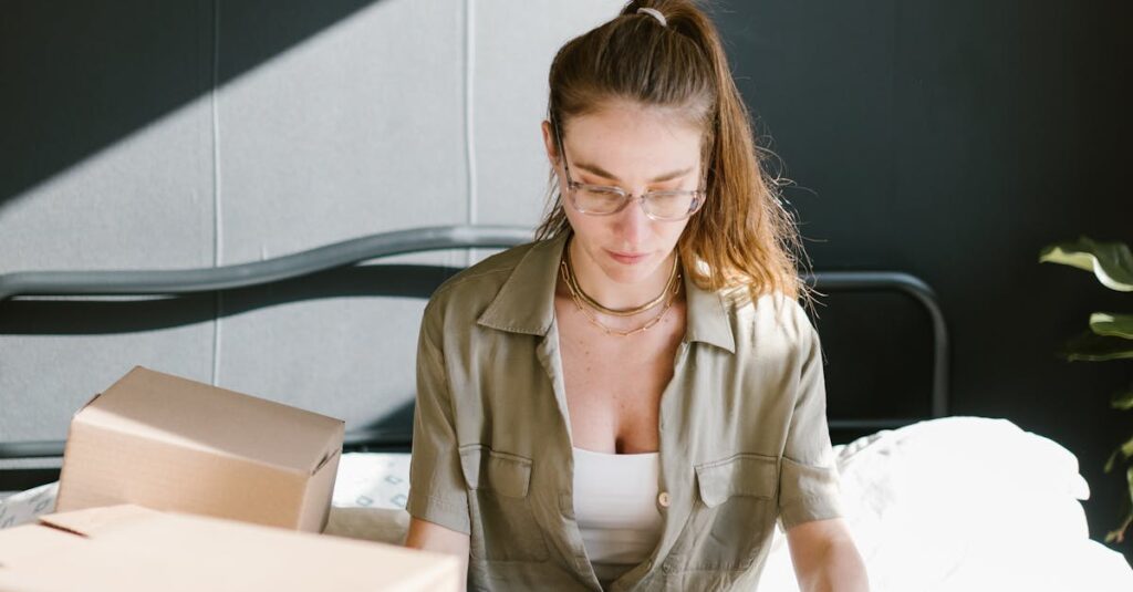 Woman sitting on a bed working on a laptop surrounded by cardboard boxes, sunny room.