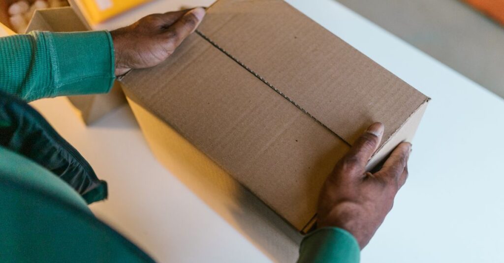 Close-up of hands holding a brown cardboard box on a white table indoors.