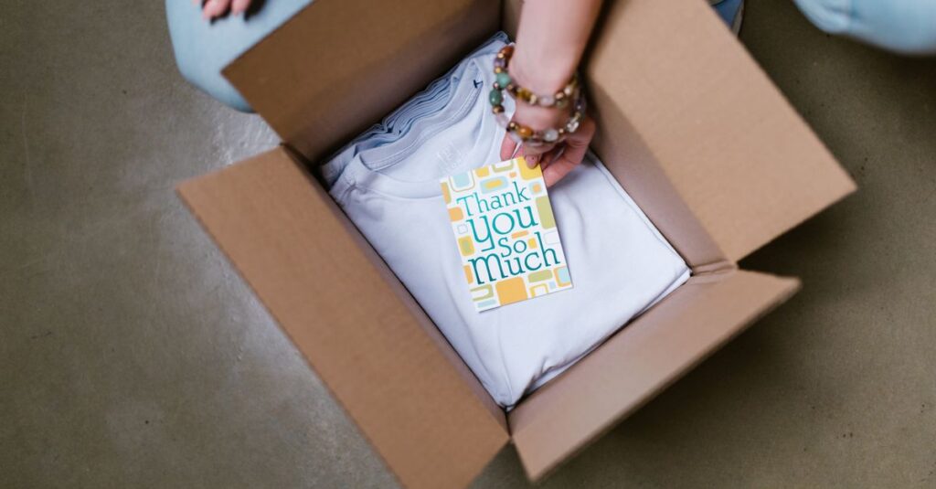 A person packing a white t-shirt with a thank you card into a cardboard box.