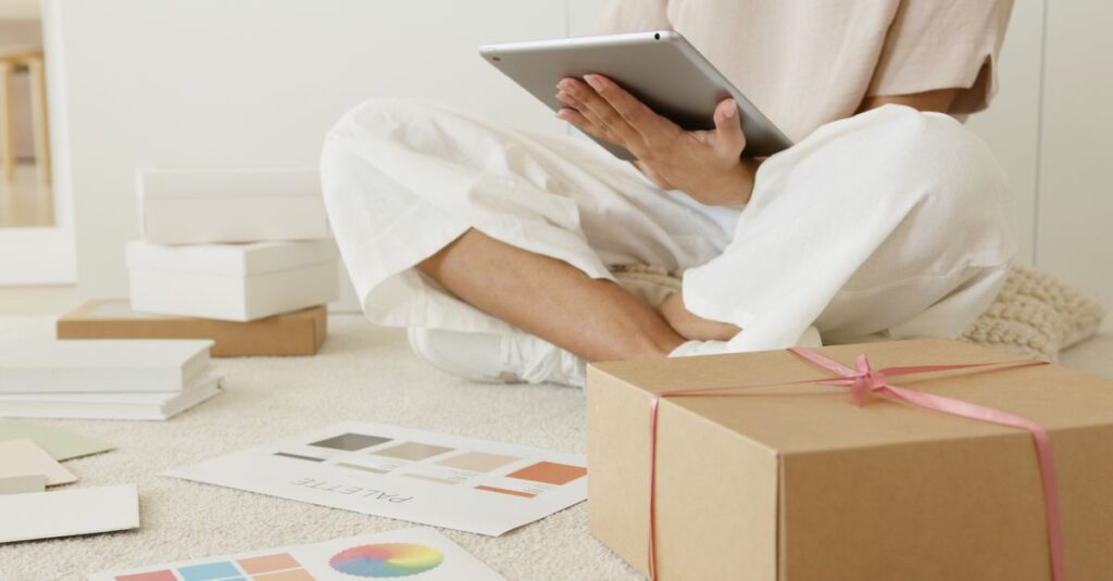 A person sitting with a tablet, surrounded by gift boxes and color palettes, in a cozy indoor setting.
