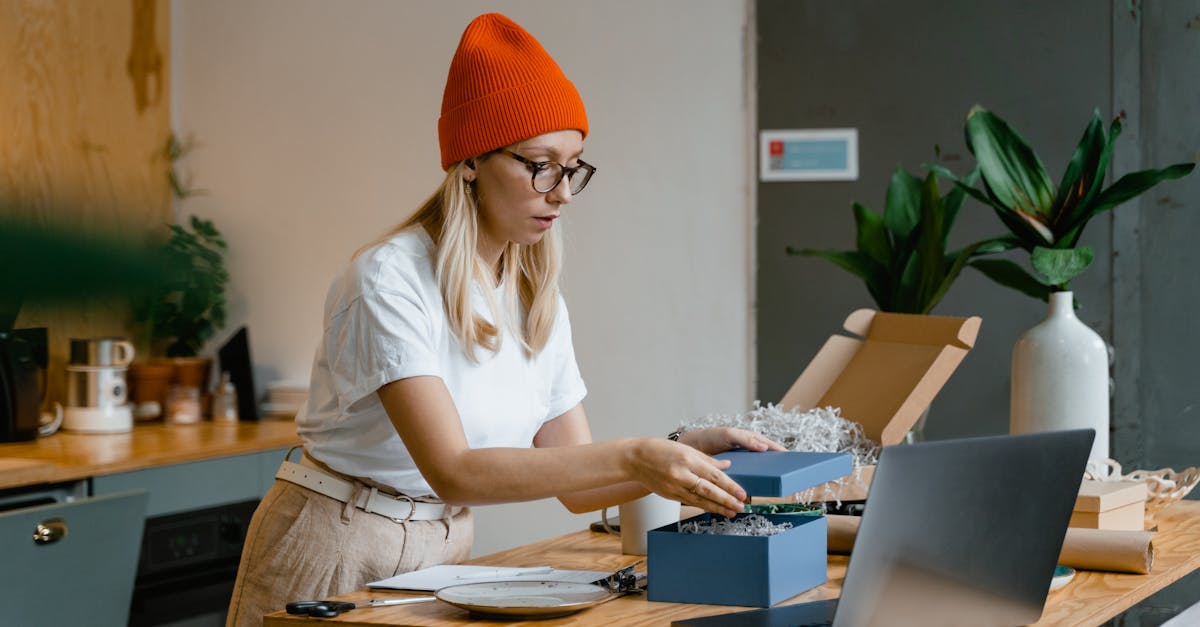 Woman packing a gift box with care at a cozy indoor workspace, using a laptop for guidance.