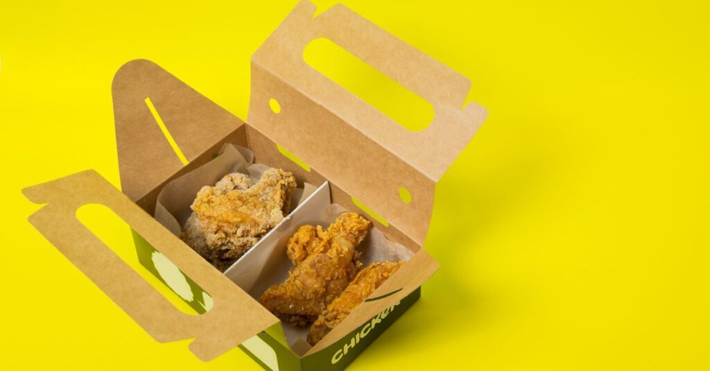 From above of opened cardboard box with takeaway fried chicken meat placed on bright yellow surface in studio