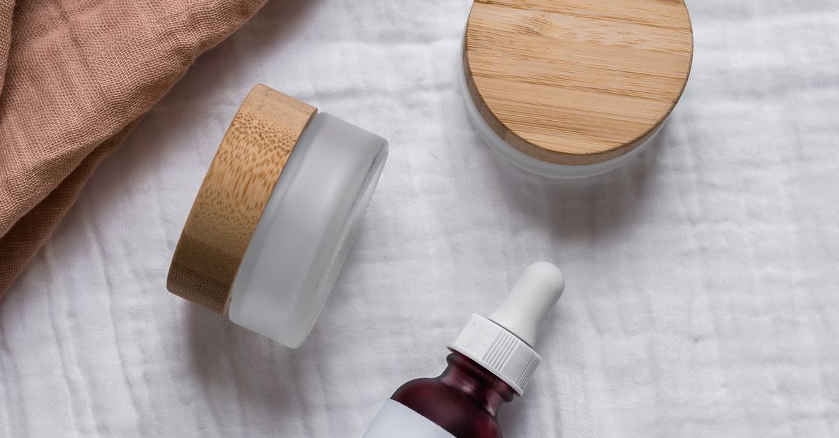 Flatlay of natural skincare jars and dropper bottle with wooden lids on a soft fabric background.