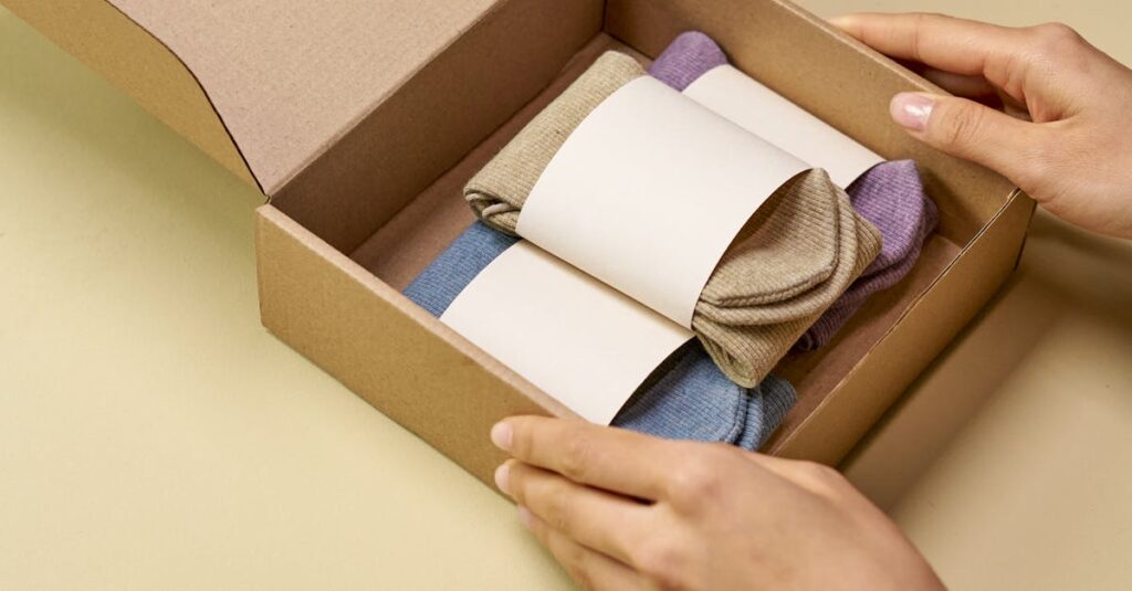 A woman's hands packing colorful socks into a cardboard box on a light yellow surface.