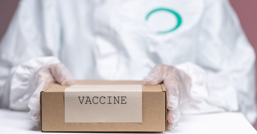 Healthcare worker holding a vaccine package in protective gear, emphasizing safety and health.